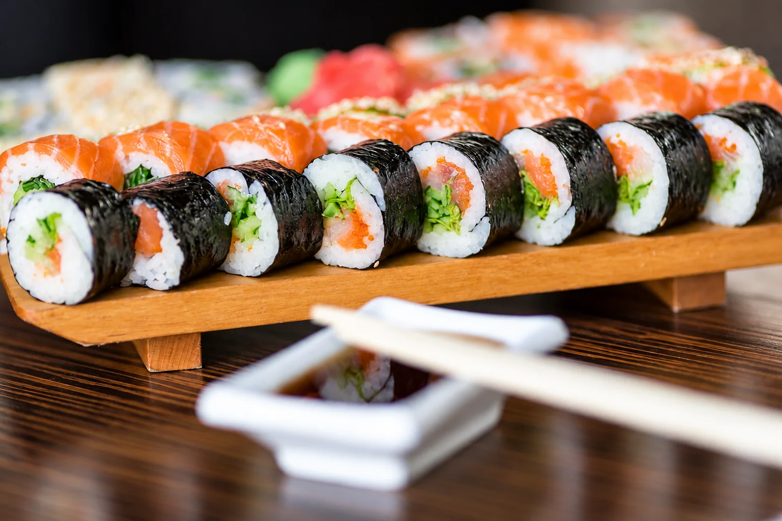 Makis alignés sur un support en bois, coupelle de sauce et baguettes au premier plan.