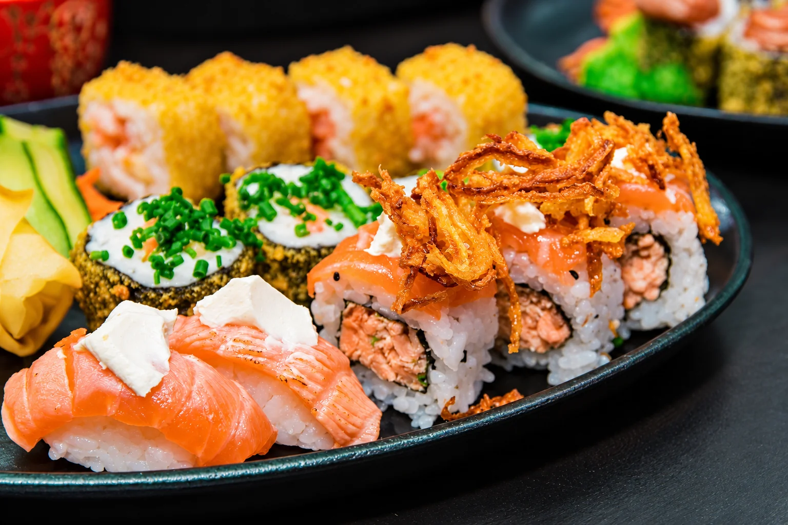 Sushis saumon, makis et rolls garnis d'oignons frits sur une assiette noire.